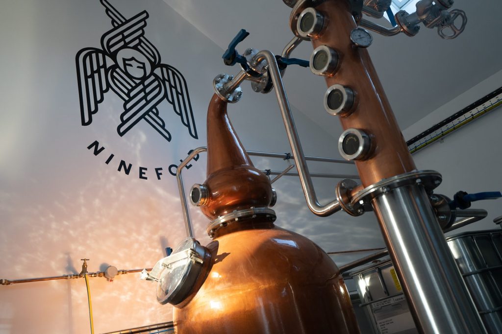 A copper pot still, with the six winged seraphim of the distillery logo on the wall behind