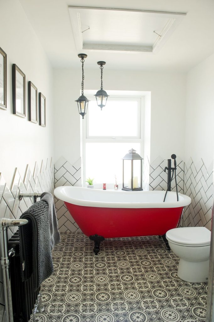 A red roll top bath beneath a window, with herringbone wall tiles and a patterned tiled floor